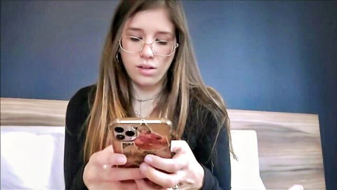 Skinny brunette teen with glasses intently looks at phone on bed