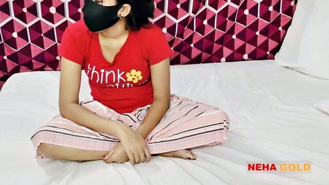 Sexy masked Indian girl Neha Gold in tight red V-neck top and pink striped pants sitting on bed