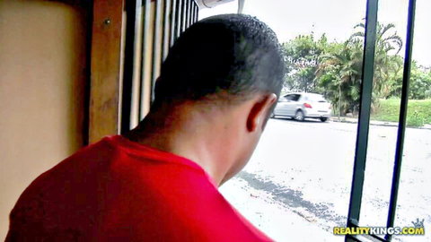 Black guy in red shirt peeking through barred window at parked silver car outdoors