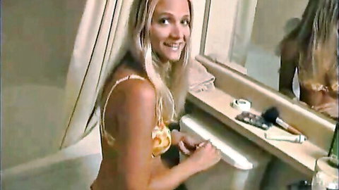 Blonde babe in yellow bikini top smiling in bathroom mirror at vanity sink
