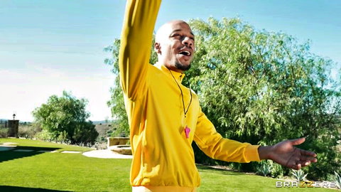 Bald Black man in yellow tracksuit raises arms excitedly outdoors on green lawn near pool
