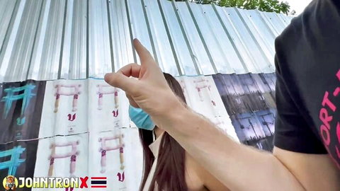 Busty smiling Asian woman exposes huge bare tits outdoors as man's finger points at her nipple by graffiti wall