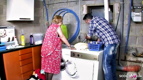 Mature blonde housewife in pink floral dress leans over washer with young man in laundry room