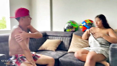 Busty topless Asian woman flashing huge tits to shirtless guy in red cap on living room couch