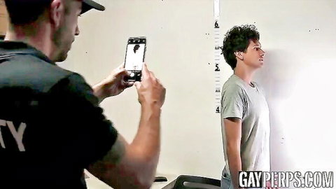 Tattooed cop in black uniform mugshots curly-haired twink suspect against police height wall