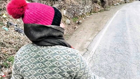 South Asian woman in pink pom-pom beanie and patterned sweater stands by stone wall on outdoor road