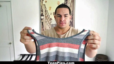 Tanned muscular man holding open gray red striped tank top in bedroom