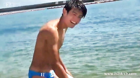 Muscular Asian hunk smiling with huge cock bulge in tight blue speedo at beach.