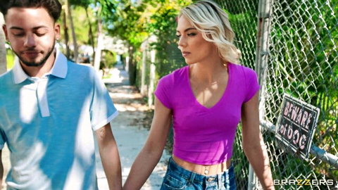 Blonde woman in pink top and jeans walks hand-in-hand with man outdoors near fence