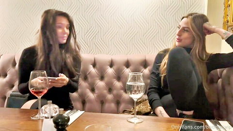 Two sexy clothed women sipping wine on pink velvet couch holding phones
