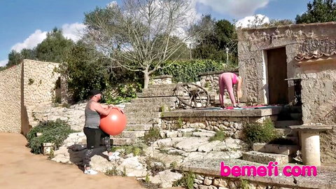 Muscular guy holds exercise ball as sexy woman bends over showing tight pink yoga pants ass outdoors