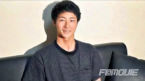 Smiling Asian man in casual t-shirt sits on couch, hinting at kinky solo uniform play