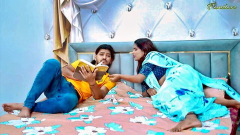 Sexy Indian couple cuddles passionately on bed, wife in blue saree leans over boyfriend reading book