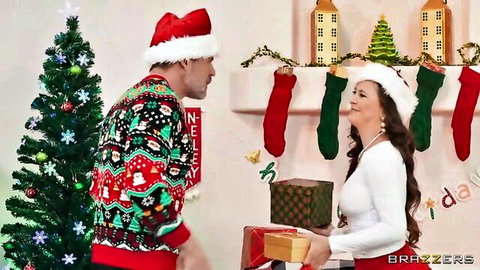 Curvy brunette MILF in Santa hat and white sweater holds Christmas gift smiling with man in ugly sweater by tree