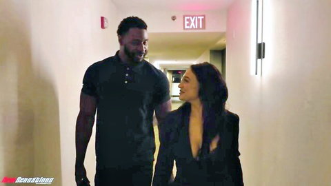 Brunette with deep cleavage blazer holds hands with muscular black stud in steamy hotel hallway