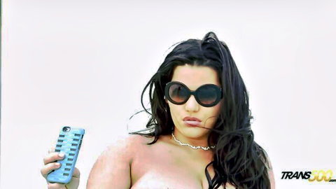 Busty oiled trans babe with huge tits poses topless holding phone remote in big sunglasses