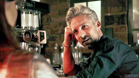 Muscular blonde man with fair skin leans at bar counter eyeing brunette long-haired woman