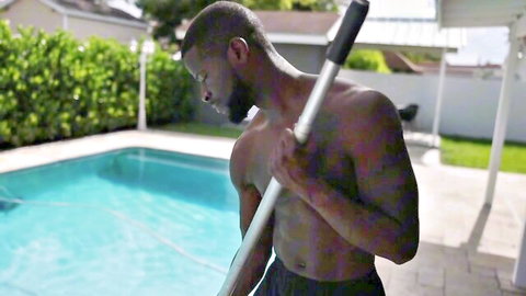 Muscular black stud shirtless in shorts holds pool skimmer by backyard pool