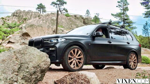 Black BMW X7 SUV parked on rocky outdoor terrain with pine trees and Vixen.com watermark
