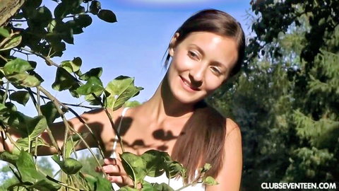 Smiling petite teen peeks playfully through green leaves in sunny outdoor forest
