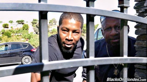 Two muscular black men peering intensely through metal fence in outdoor interracial teaser