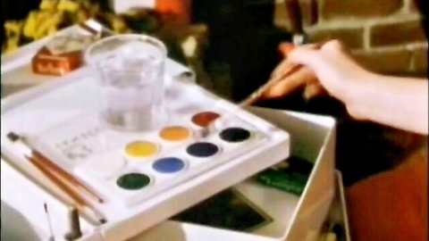 Hand dipping paintbrush into red paint on watercolor palette with water glass nearby