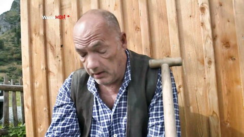 Egon Kowalski mature hunk in plaid shirt and vest, bald with mustache, holding wooden staff outdoors