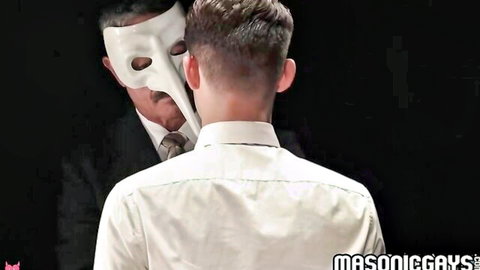 Masked man in white phantom mask leans close behind young suited twink in gay tease scene