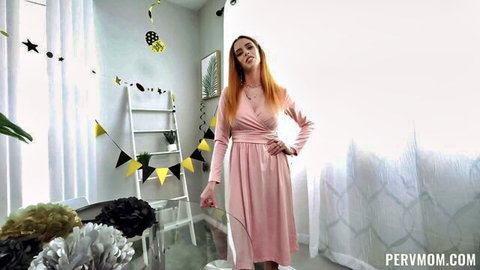 Redhead MILF in pink wrap dress posing seductively with hands on hips in party room