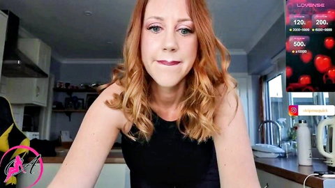 Sexy redhead cam girl smiles teasingly in black top on kitchen webcam with Lovense