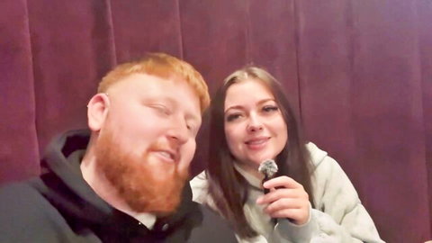 Red-bearded guy and brunette babe smiling close together holding mic in steamy purple booth