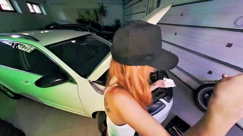 Sexy redhead in tight white tank top bends over open car hood in garage showing curves