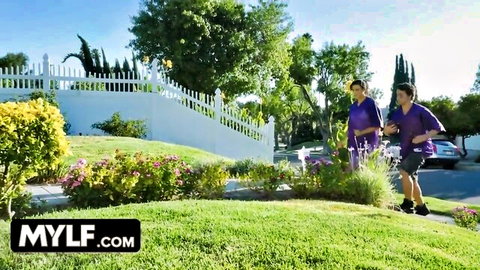 Brunette MILF in purple top walks with muscular stepson in purple shirt on sunny suburban lawn