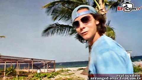 Handsome long-haired guy in sunglasses and tie-dye shirt smiles on beach for real sexdate pickup