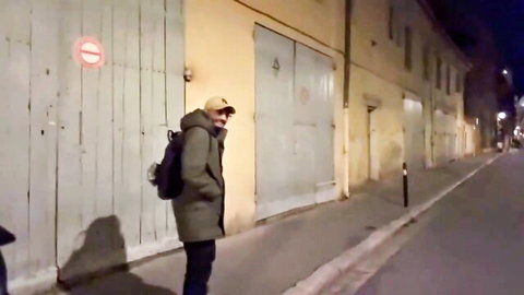 Man in yellow cap and coat with backpack stands on dark French alley street at night