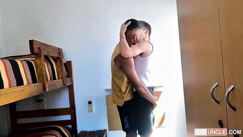 Muscular guy in tank top passionately hugs shirtless hunk from behind in bunk bed dorm room