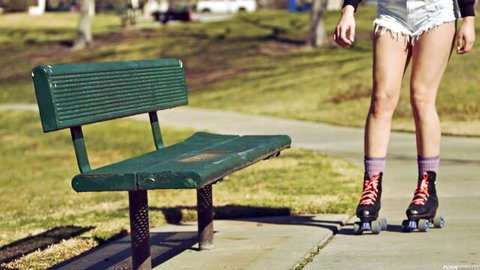 Skinny blonde teen in tiny white shorts and red roller skates teasing beside green park bench