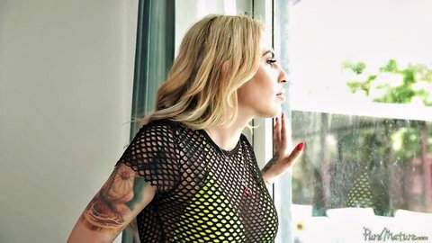 Tattooed blonde babe in sheer fishnet top teasing curves by window