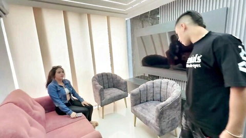 Sexy Asian teen in denim jacket and bralette sits teasingly on pink sofa with handsome guy standing by