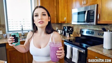 Lily Lou in white tank top holding green can and purple smoothie, sexy kitchen pose