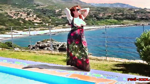 Busty redhead in floral dress poses arms up by pool with sea view