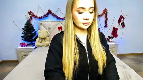 Cute young blonde in black hoodie smiles on Christmas webcam bed.