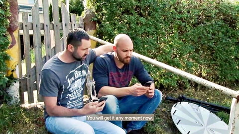 Two bearded men in jeans and t-shirts sitting outdoors holding phones casually