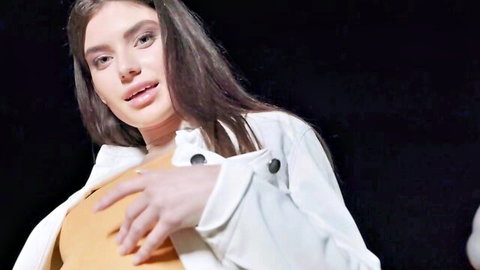 Seductive long-haired brunette in open white jacket teasing her cleavage with hand on orange top