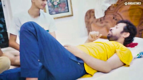 Bearded Indian DILF lying on bed pants lowered young guy in white shirt stands chatting bedroom tease