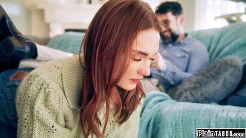 Redhead woman in green sweater leans intimately toward smiling man on couch in taboo home scene