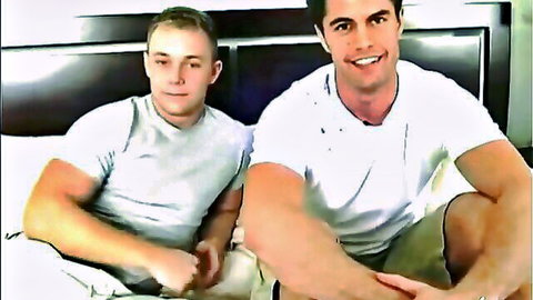 Two muscular gay hunks in tight white shirts smiling seductively on bed teasing gay action
