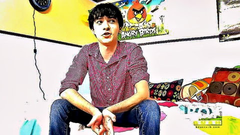 Cute young man with curly hair sits clothed on bed in colorful bedroom with Angry Birds posters