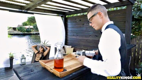 Tanned bikini babe with small tits lounges seductively as handsome waiter serves whiskey at outdoor bar