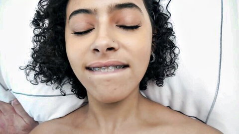 Braces teen Latina with curly hair lies in bed eyes closed smirking with open mouth teasing oral